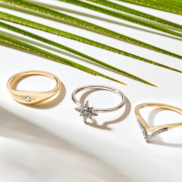 Three rings displayed on a white surface with green palm leaves in the background: a gold ring with a small star motif, a silver ring with a starburst center, and a gold ring with a V-shaped pavé band.