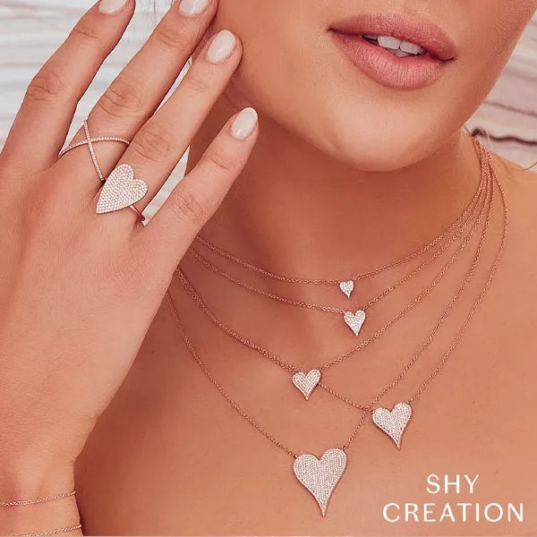 A close-up lifestyle shot of a model wearing rose gold jewelry, featuring a multi-layered necklace with heart pendants and a matching heart ring on her finger. The brand name 'SHY CREATION' is visible in the bottom right corner.