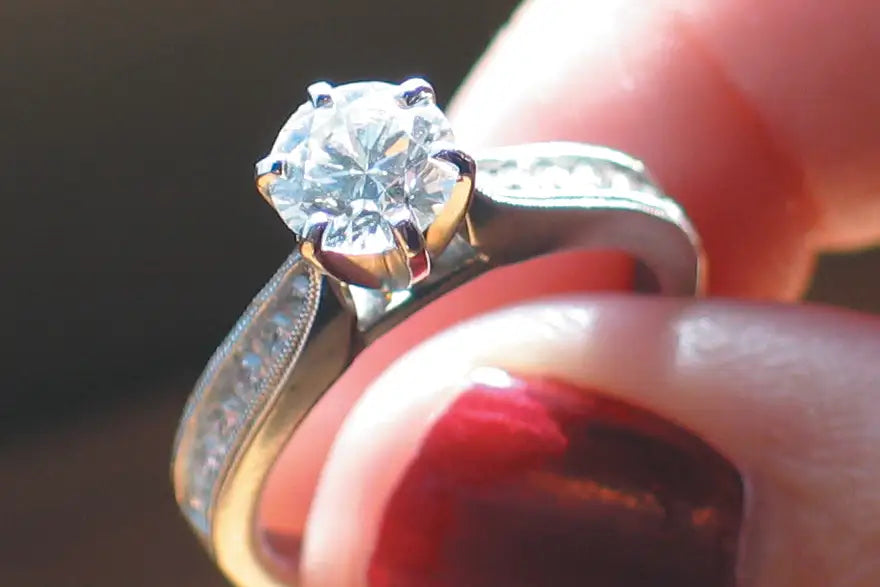 A close-up shot of a diamond engagement ring held between fingers. The ring features a round brilliant-cut diamond set in a yellow gold or gold-plated four-prong setting, with a silver-colored pavé band.