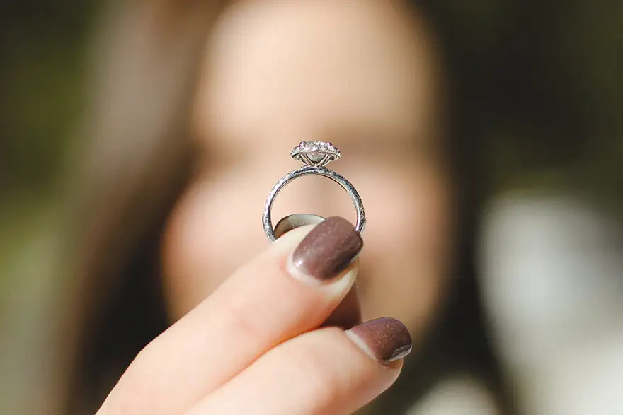 A silver-toned diamond ring held between fingers with brown nail polish, against a heavily blurred background of a person's face.
