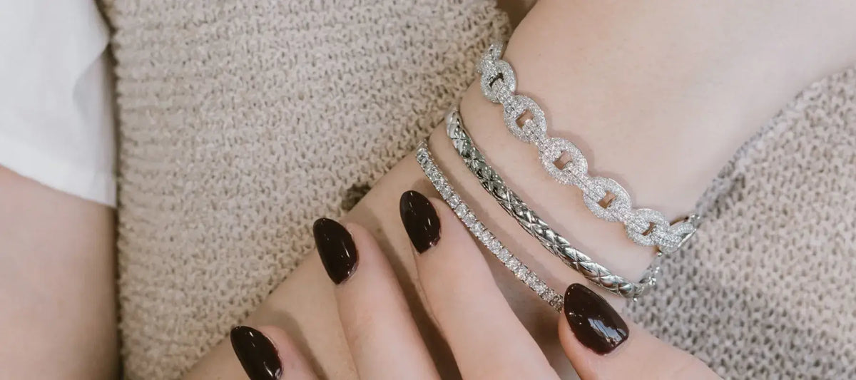 A close-up of a person's wrist and hand wearing three silver chain bracelets, including a link chain bracelet, a twisted rope bracelet, and a diamond-encrusted bracelet, against a beige knit sweater.