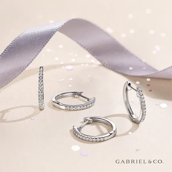 A set of silver huggie hoop earrings with pavé-set clear stones, arranged on a light beige surface with a lavender satin ribbon and confetti. The brand name GABRIEL & CO.