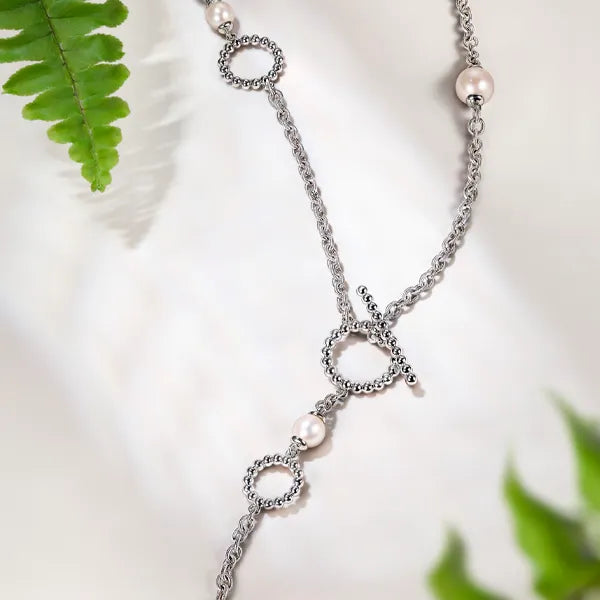 A silver chain necklace with three textured circular ring charms, each accented with a small white pearl bead, displayed on a soft white fabric background with green fern leaves.