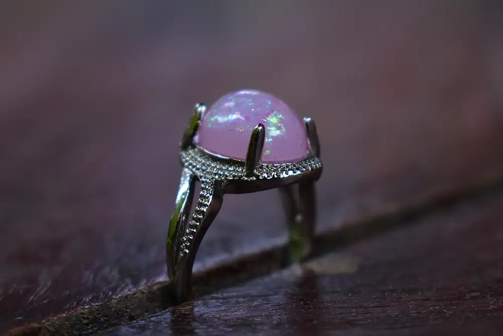 A silver ring featuring a large, oval, iridescent light pink cabochon gemstone, set on a textured dark brown wooden surface.