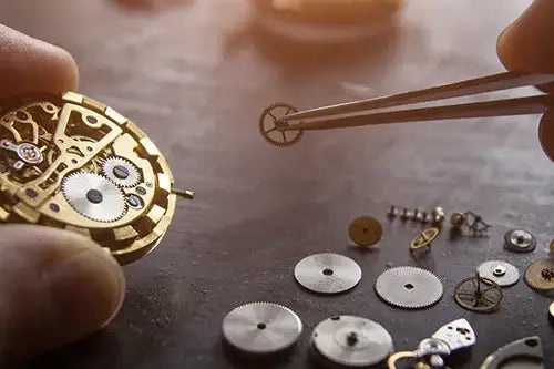 Close-up of a watchmaker's hands assembling a gold-toned mechanical watch movement, using tweezers to place a small gear onto a workbench surrounded by watch parts.