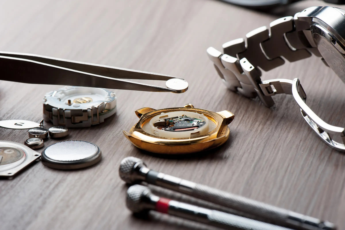 A close-up of a disassembled gold-tone watch with its metal bracelet, tweezers, tools, and small watch parts arranged on a wooden surface.