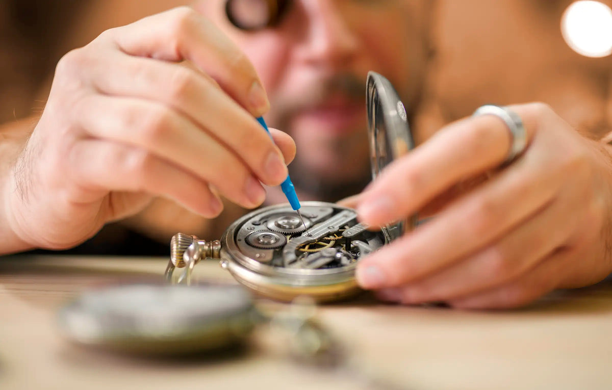 A close-up of a person's hands disassembling and examining the internal mechanism of an open pocket watch on a wooden workbench.