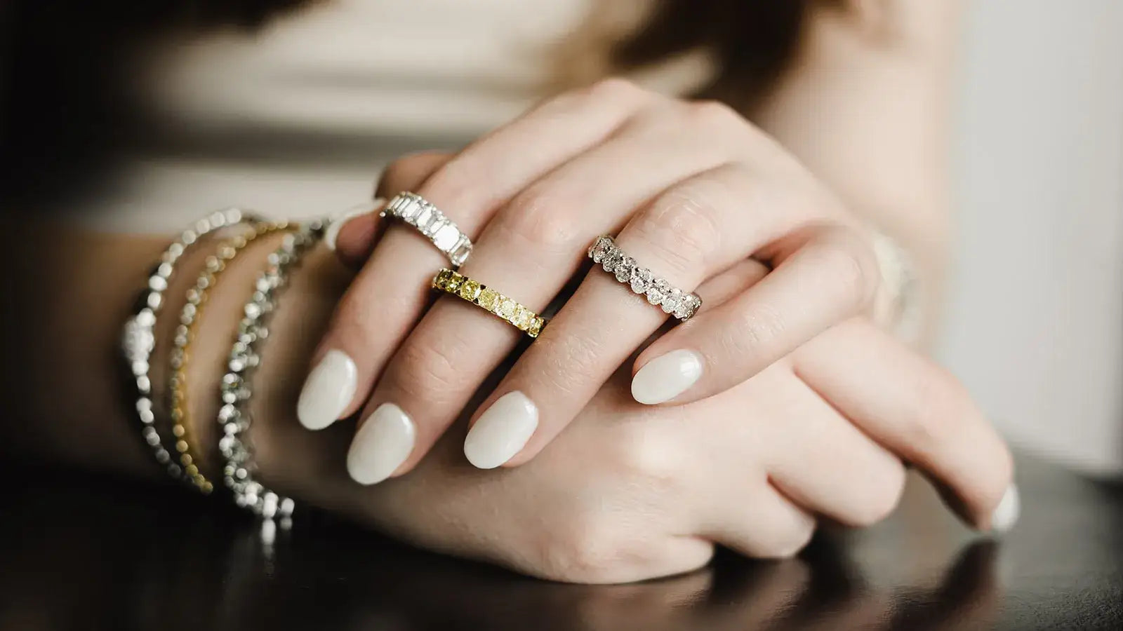 A close-up of a person's hands resting on a dark surface, wearing several rings and bracelets. The rings include silver and yellow gold bands with clear gemstones, and the hands have white manicured nails.